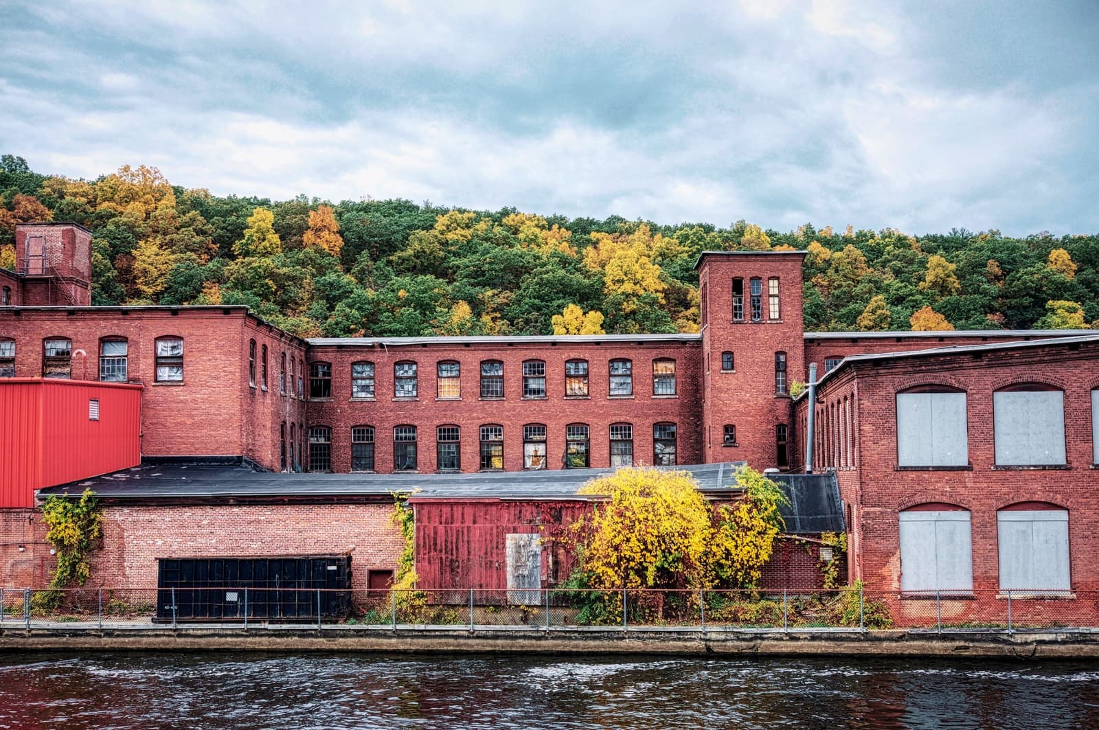 Red-brick textile mill on a river with autumn hills behind, like the mill lofts movers in Woonsocket, RI empty and fill every month